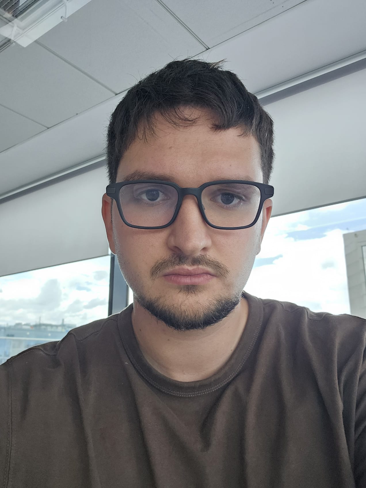 A selfie of a young man with glasses and a short beard, taken in an office in front of a window.