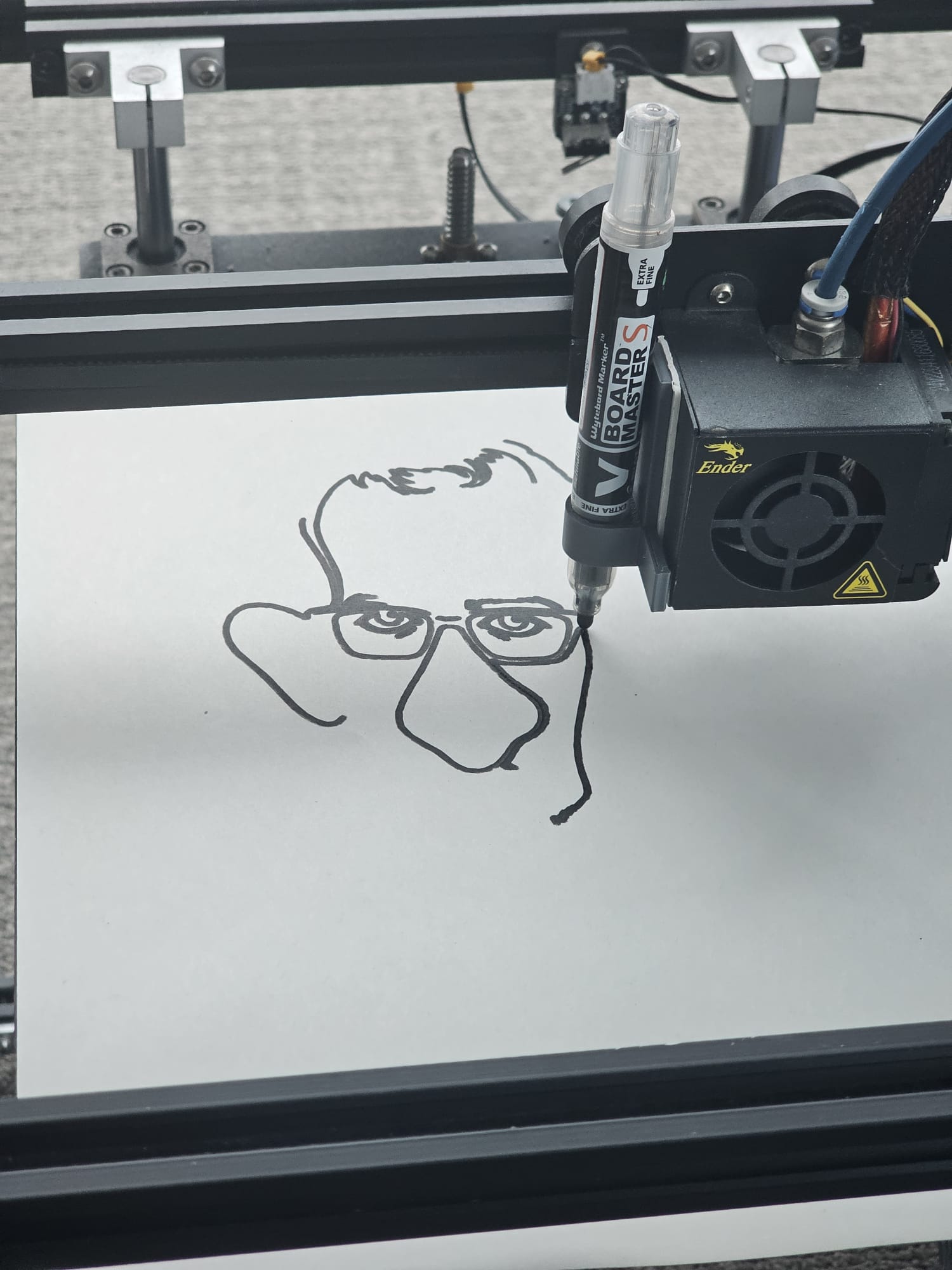 An Ender 5 Pro 3D printer partway through a drawing, a whiteboard marker clamped to the print head next to the hot end, drawing a line-art caricature of a face with glasses on white paper.